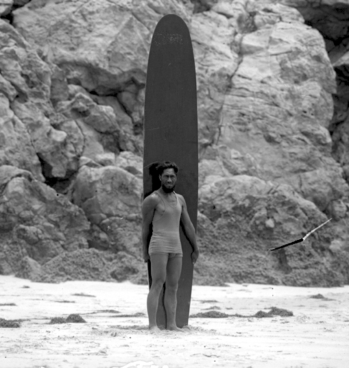 Photo du surfeur et champion olympique de natation Duke Kahanamoku debout devant sa planche de surf sur une plage de Los Angeles, en Californie, vers 1920 