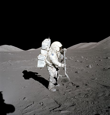 Shown is a black and white photograph of a person in a spacesuit, using a rake on grey, dusty ground.