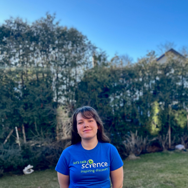 Julia se tient dans un champ sur fond de feuillage vert et de ciel bleu vif. Elle porte un t-shirt bleu Let's Talk Science, ses cheveux bruns tombent sur ses épaules et elle sourit à l'appareil photo.