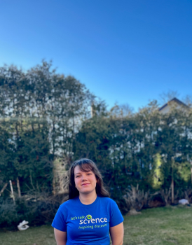 Julia se tient dans un champ sur fond de feuillage vert et de ciel bleu vif. Elle porte un t-shirt bleu Let's Talk Science, ses cheveux bruns tombent sur ses épaules et elle sourit à l'appareil photo.
