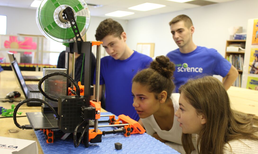 Trois élèves observent attentivement une imprimante 3D, sous le regard d’un bénévole de Parlons sciences.