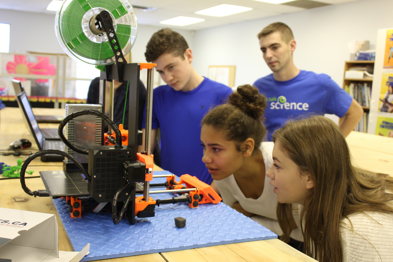 Trois élèves observent attentivement une imprimante 3D, sous le regard d’un bénévole de Parlons sciences.