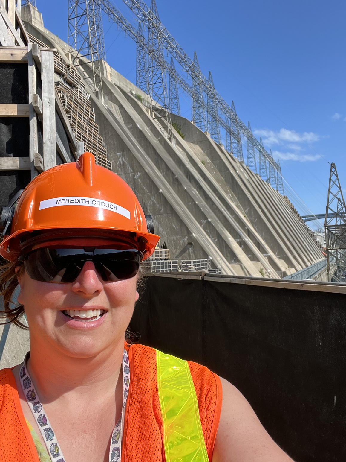 Autoportrait de Meredith Crouch portant un casque de protection et un gilet réfléchissant orange avec un barrage hydroélectrique et des fils de transmission en arrière-plan
