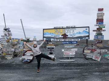 Charlotte Cockburn standing in front of signs indicating distance to a variety of locations from Alert, Nunavut.   