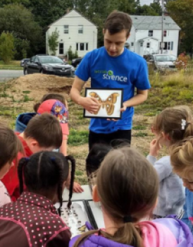  Des élèves du primaire rassemblés à l’extérieur avec un bénévole vêtu d’un t-shirt bleu Parlons sciences qui montre le dessin coloré d’un papillon. 